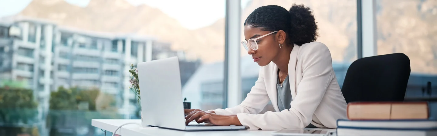 A woman in glasses and white blazer works on a laptop at a desk with mountains and buildings visible through the window.