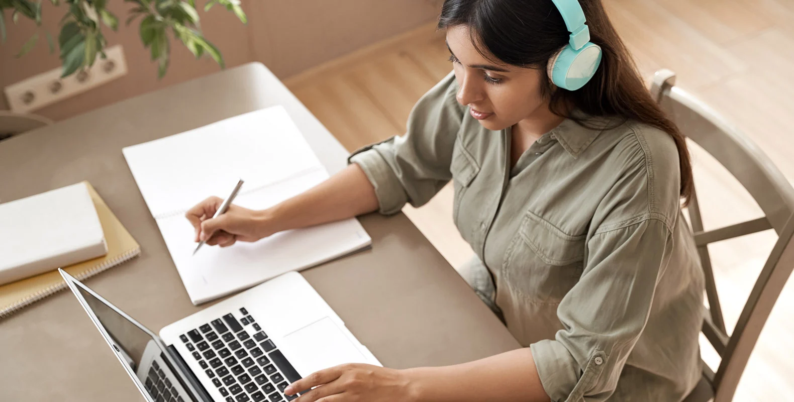 A person with headphones writes in a notebook while using a laptop at a desk.