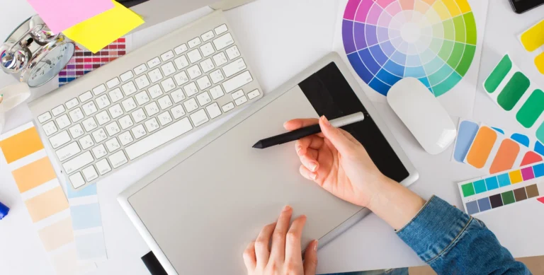 Person using a graphic tablet with a stylus, surrounded by a keyboard, color swatches, and a color wheel on a desk.