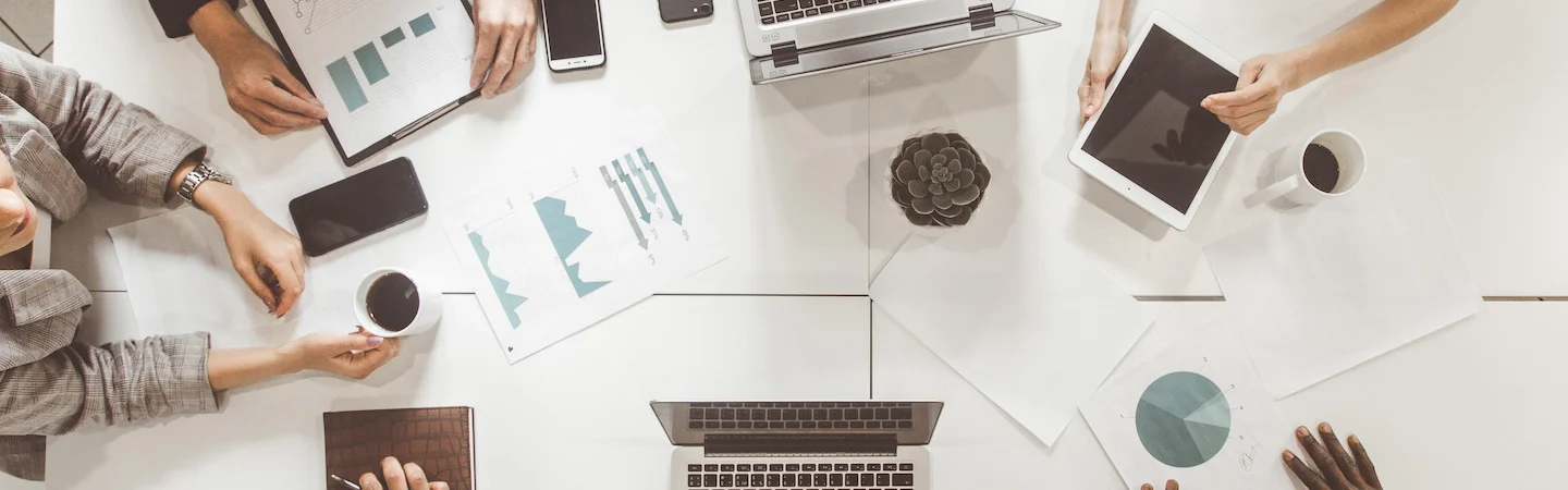 A top view of a meeting table with laptops, tablets, documents, and cups of coffee. Four people are seen collaborating.