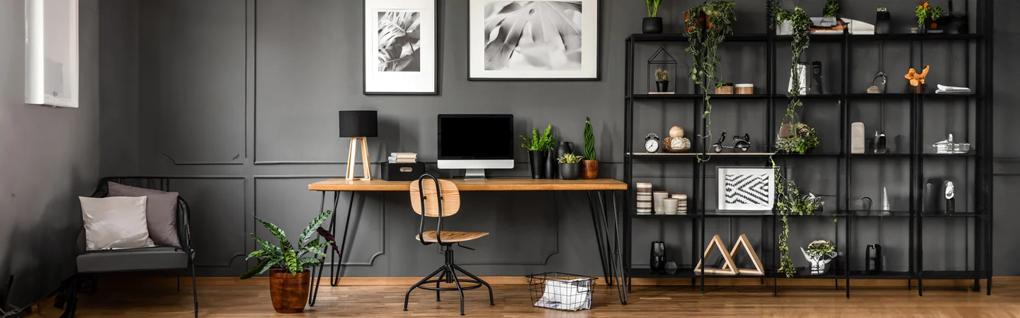 Modern home office with a wooden desk, chair, computer, and black shelving unit with plants and decor against a gray wall.