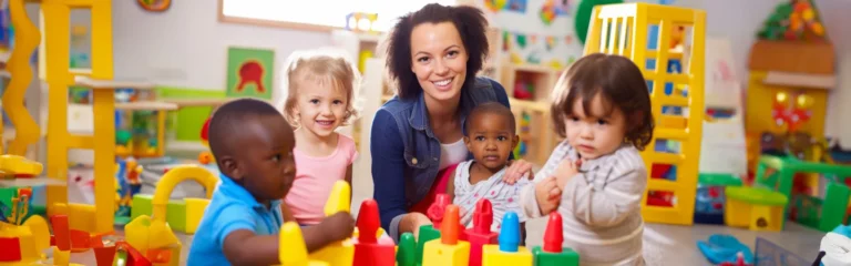 A smiling woman in a playroom surrounded by four young children playing with colorful toys.