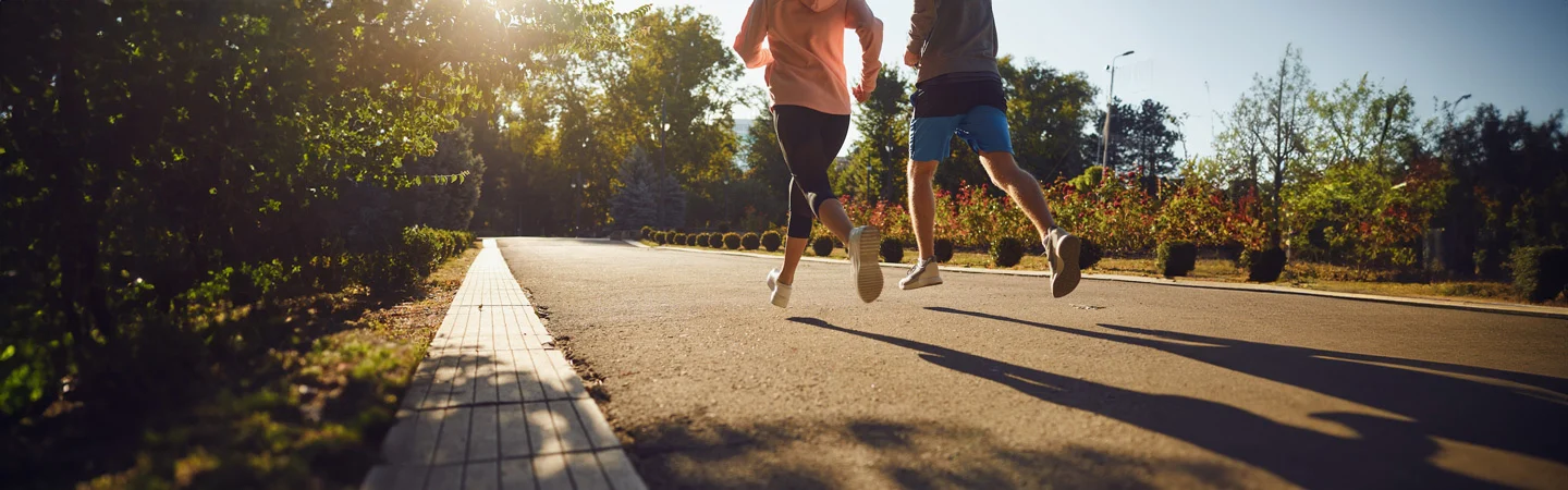 Two people jogging on a sunlit park path, surrounded by trees and greenery, casting long shadows on the ground.