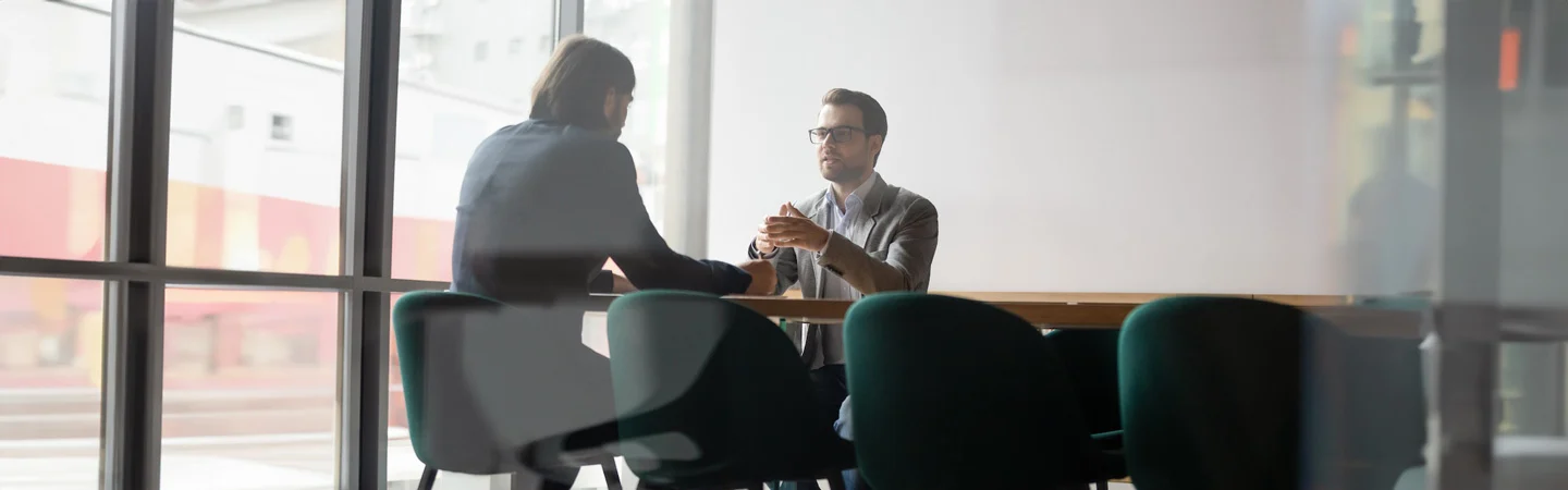 Two people are seated at a table in an office, engaged in a serious conversation next to a large window.