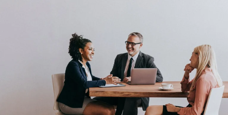 Three people in business attire having a meeting around a wooden table, with a laptop and coffee cups in front of them.