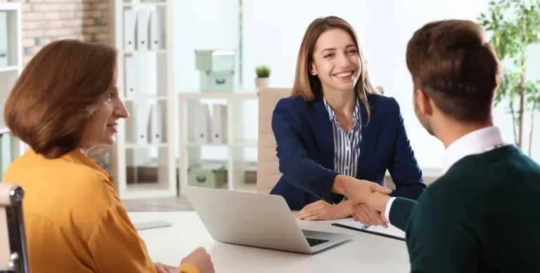 Business meeting with three people: a woman in a blue blazer shaking hands with a man, and another woman watching.