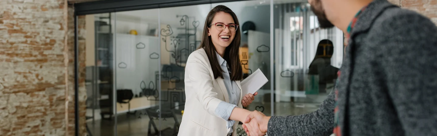 Smiling woman in a white blazer and glasses shakes hands with a man in an office with glass walls and doodles.