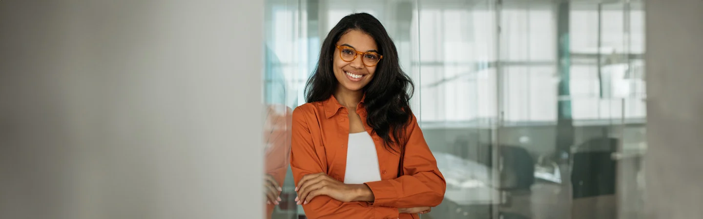 A woman in glasses and an orange shirt smiles confidently, standing with crossed arms in a modern office setting.