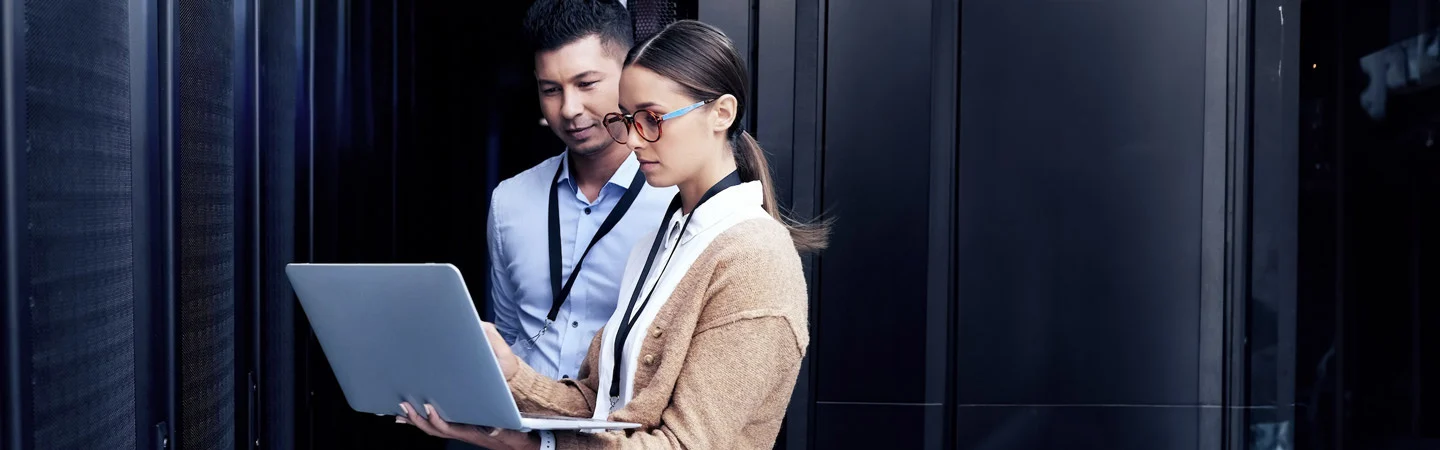 Two people standing in a server room, looking at a laptop and working collaboratively.