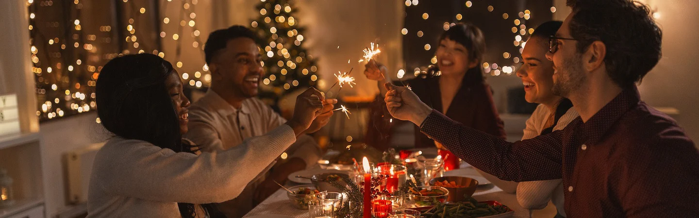 A group of people at a festive dinner table, holding sparklers with a decorated Christmas tree in the background.