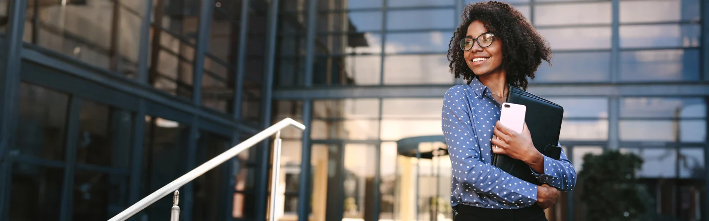 Smiling woman stands outside a modern building, holding a notebook and her phone. She wears glasses and a blue polka dot shirt.