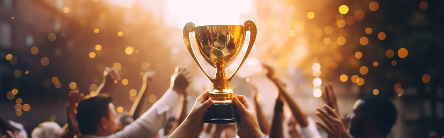 A hand holds up a golden trophy amid a cheering crowd with blurred lights in the background.