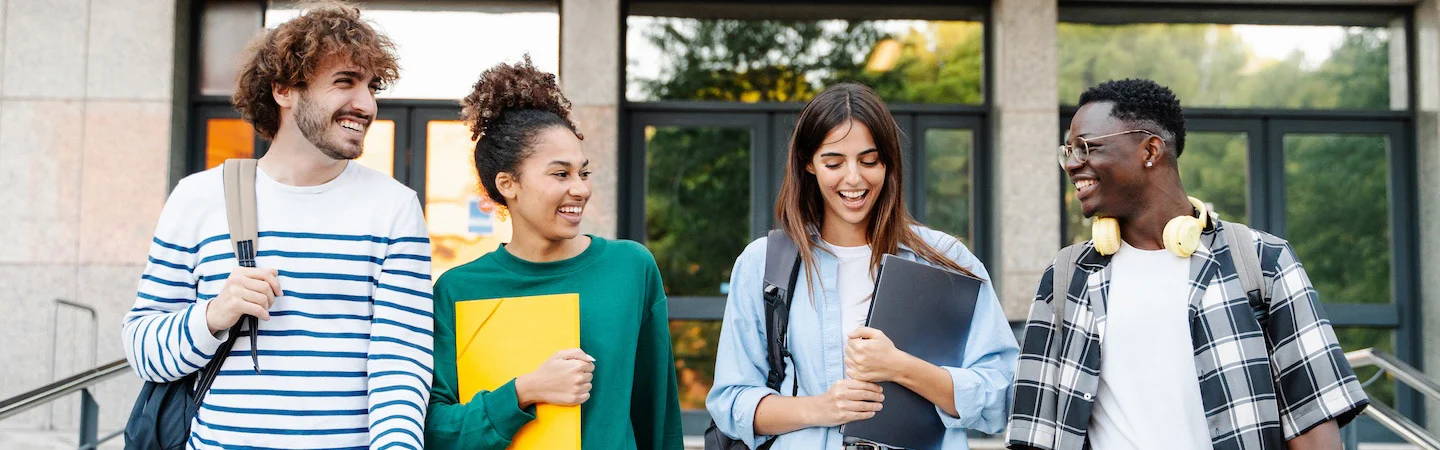 Four students carrying bags and folders, smiling and chatting while walking down steps outside a building.