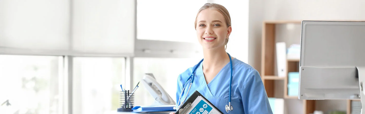 Smiling healthcare professional in blue scrubs holding a tablet in a well-lit medical office.