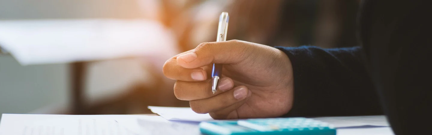 Close-up of a hand holding a pen over papers, with a calculator visible in the foreground.