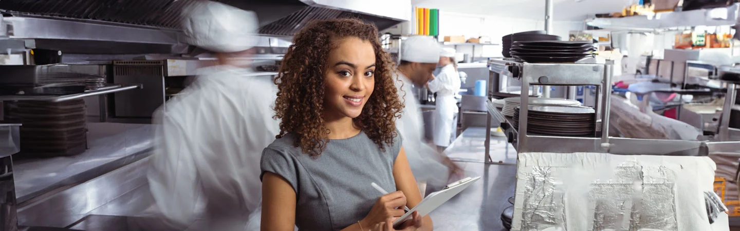 A woman with curly hair holds a clipboard, smiling in a busy commercial kitchen with chefs working in the background.