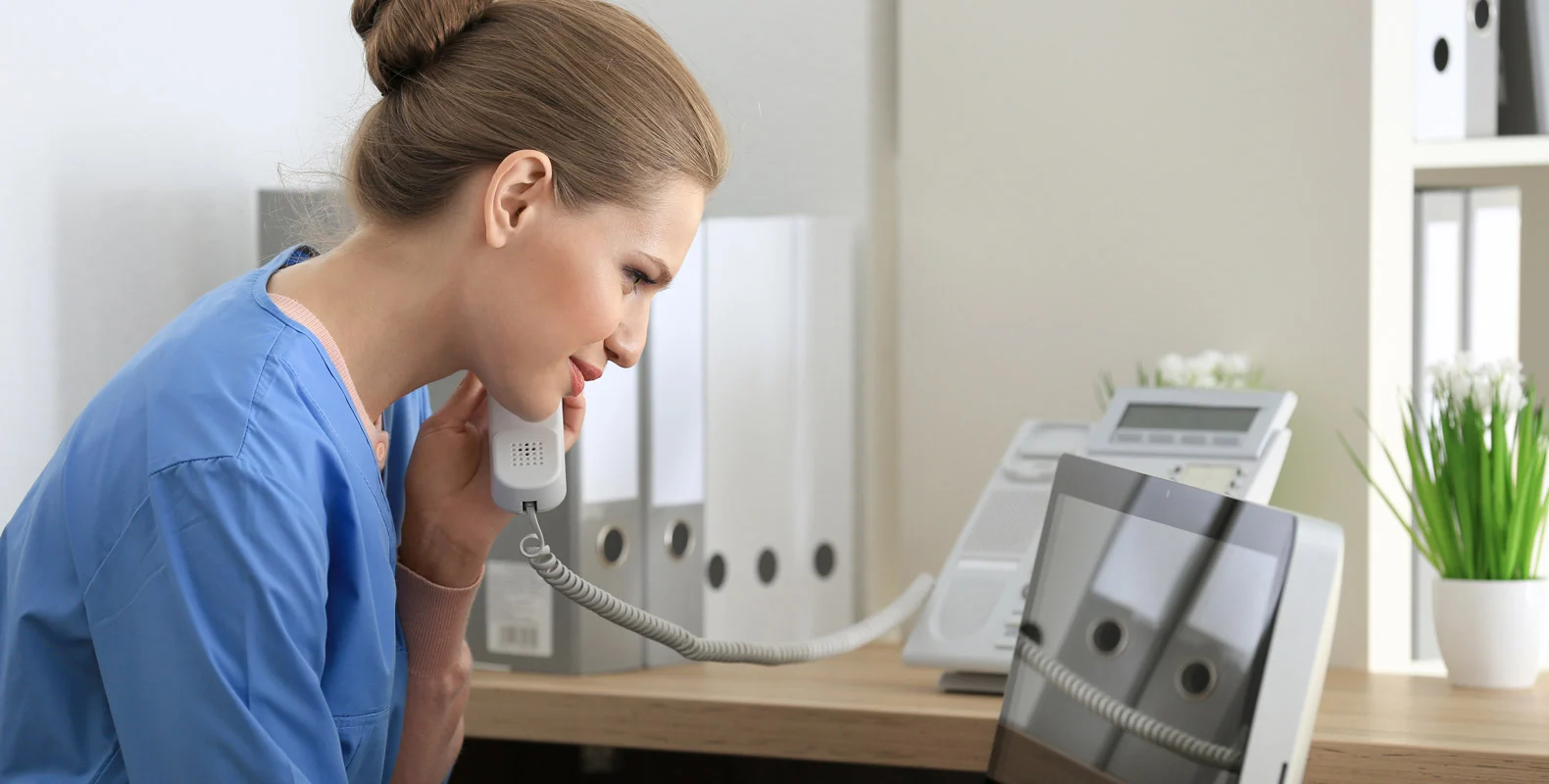 A woman in blue scrubs talks on a landline phone while looking at a computer screen in an office setting.