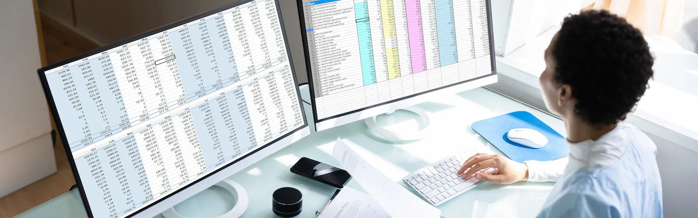 Person working at a desk with two large monitors displaying spreadsheets and colorful data charts in a bright room.