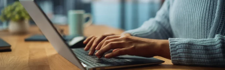 Person in a blue sweater typing on a laptop at a wooden desk with a blurred background, including a mug and plant.