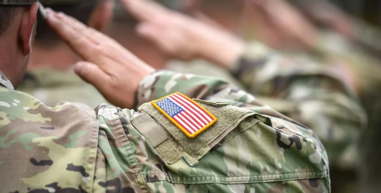 A group of soldiers in camouflage uniforms salute, with an American flag patch visible on one soldier's shoulder.