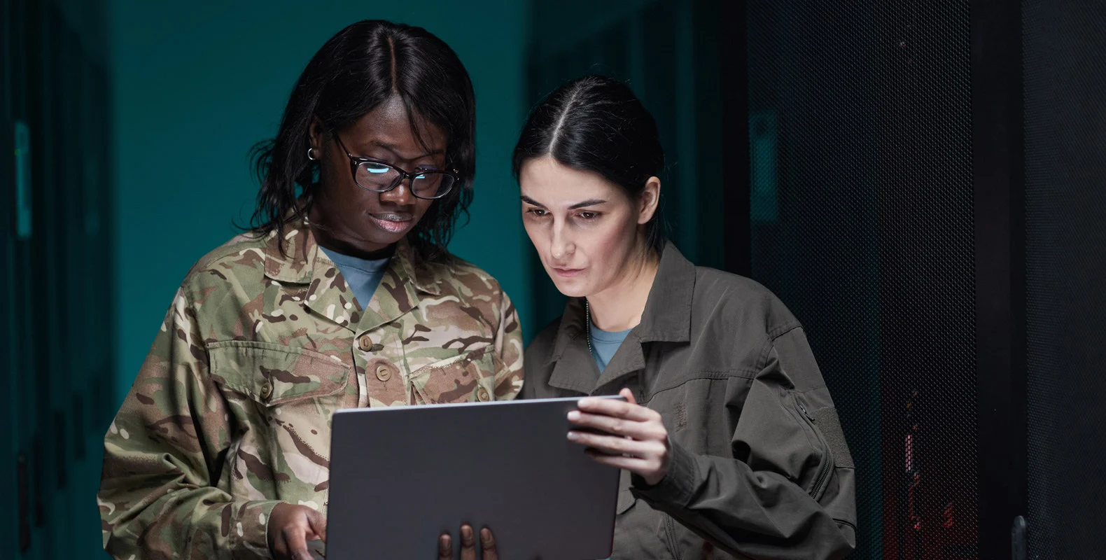 Two women in military uniforms examining a laptop in a dimly lit room.