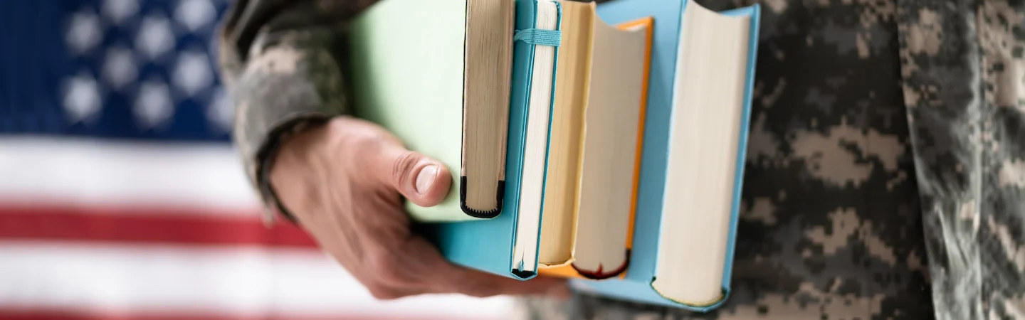 A person in military uniform holds several books in front of a blurred American flag background.