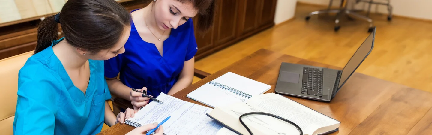 Two people in scrubs study medical notes together at a wooden table with a laptop and stethoscope.