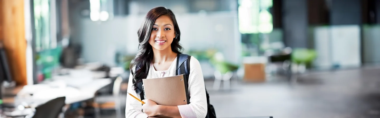 A young woman stands in a modern office holding a notebook and pencil, smiling, with desks and chairs in the background.