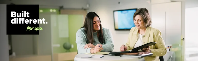 Two women in a modern office setting, one reviewing a binder and the other taking notes, with "Built different. for you." text.