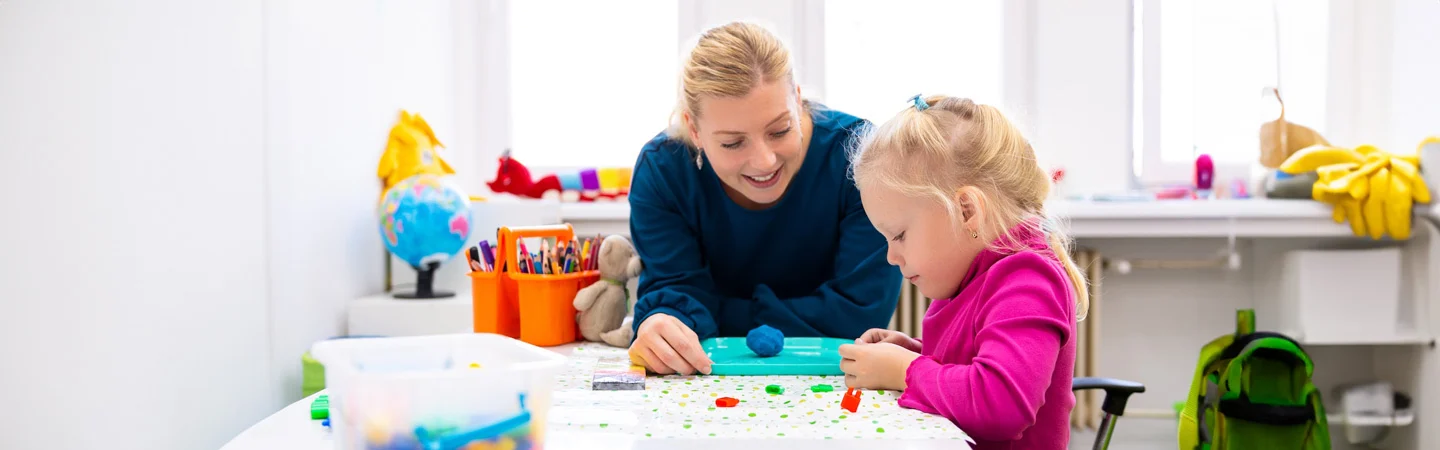 A woman assists a young girl with an art project at a table in a bright, well-organized classroom.