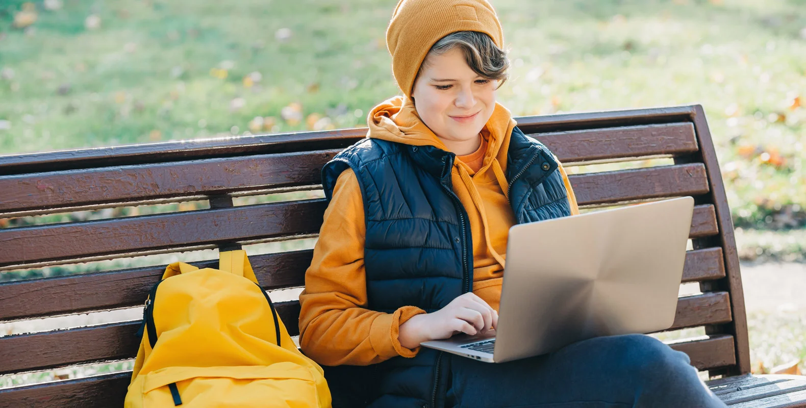 Person in yellow beanie and jacket using a laptop while sitting on a park bench with a yellow backpack beside them.