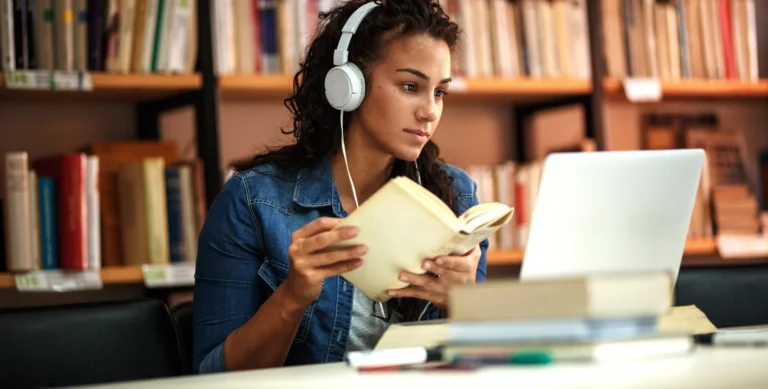 Person wearing headphones, reading a book, and looking at a laptop in a library.