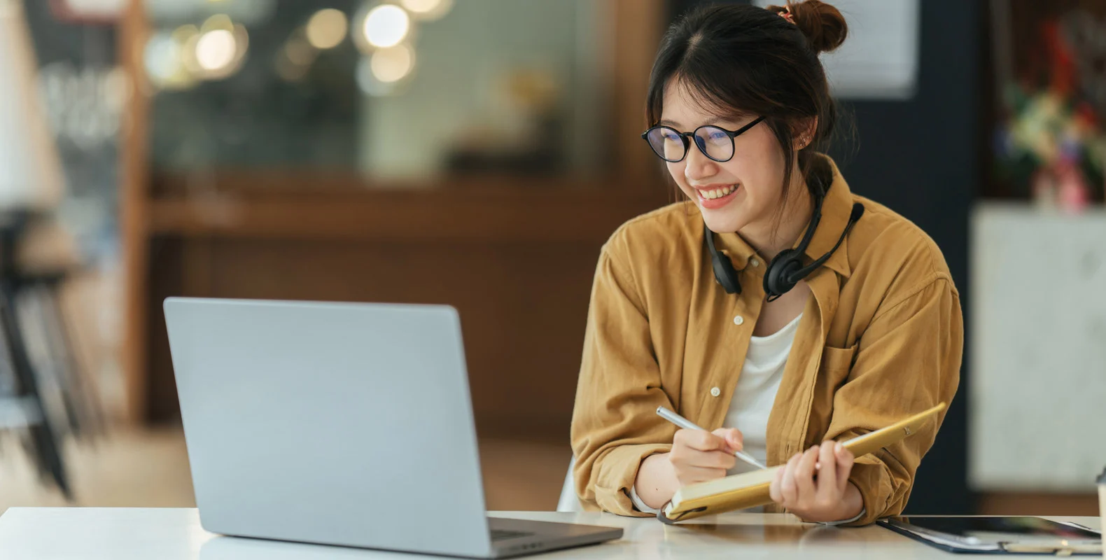 A woman wearing glasses and headphones, smiling while writing in a notebook and looking at her laptop.