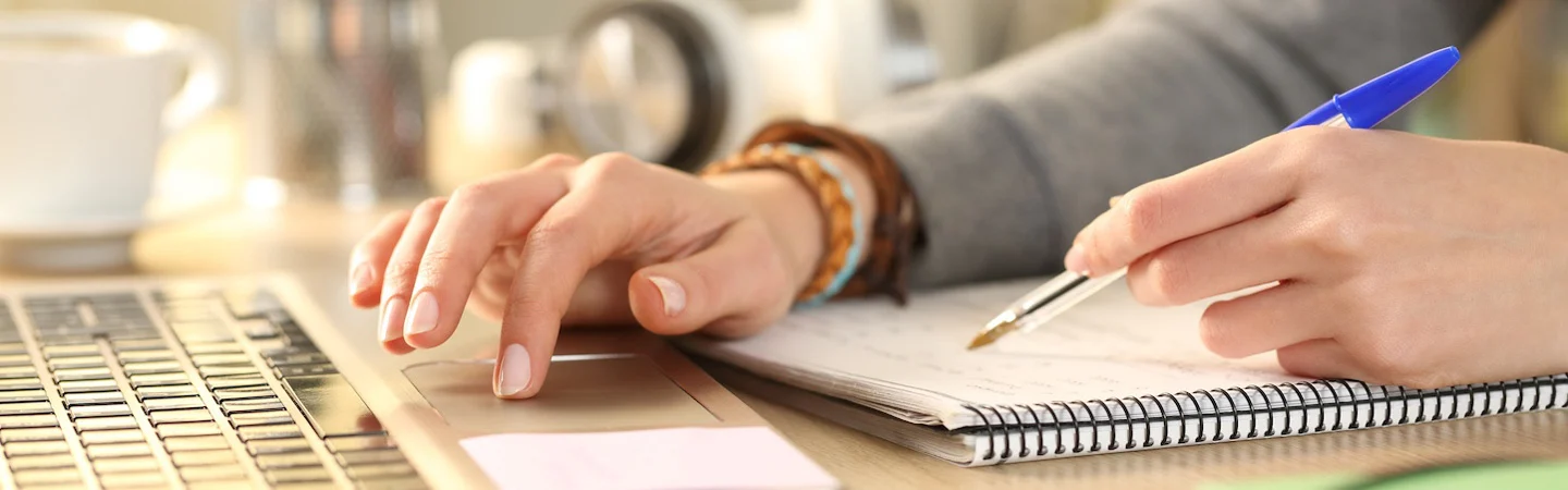 Hands of a person typing on a laptop and taking notes in a spiral notebook with a pen.