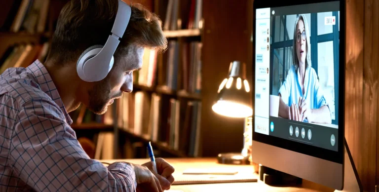 Person wearing headphones participating in a video call and taking notes on a desk with a lamp, bookshelves in background.