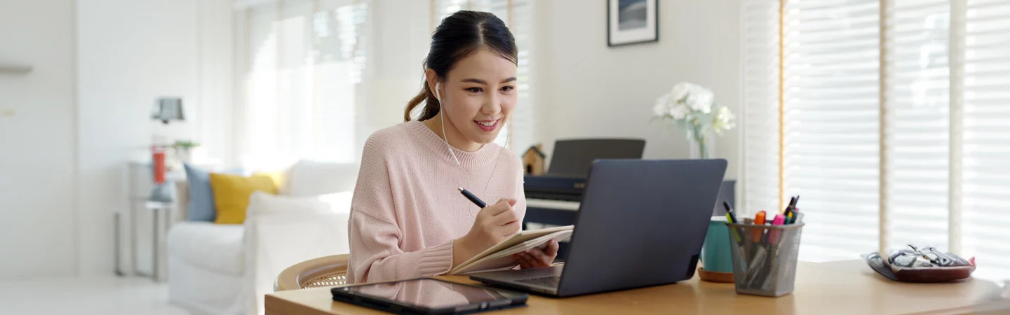 A woman smiles while taking notes and working on a laptop at a wooden desk in a bright, modern room.