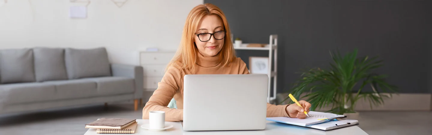 Woman with glasses working on a laptop, writing in a notebook, with a coffee cup and office items on the desk.