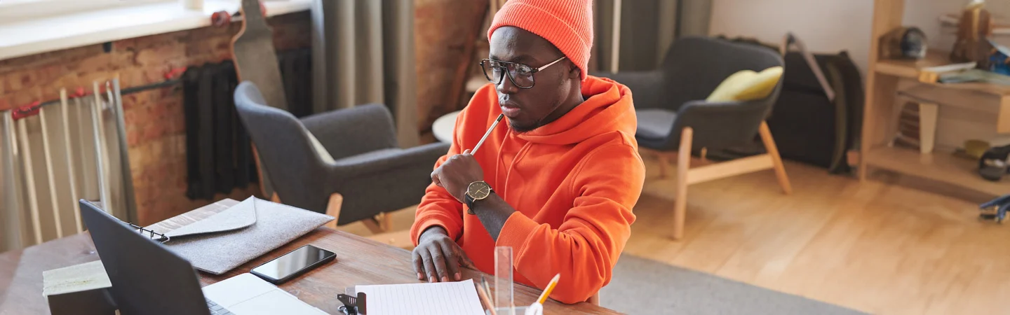 A person in an orange hoodie, wearing glasses, sits at a desk with a laptop and notepad in a cozy, modern room.