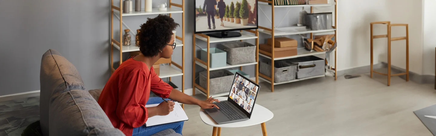 A person on a couch works on a laptop next to a notebook in a neatly organized room with shelves and a TV in the background.