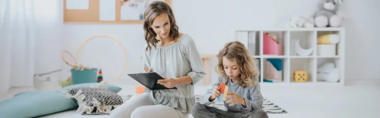 A woman takes notes on a clipboard while a child builds with toy blocks in a bright, organized room.