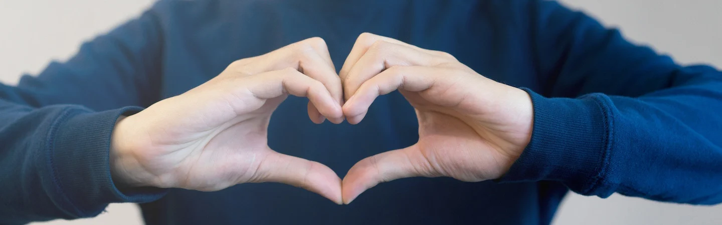 Person in a blue sweater forming a heart shape with their hands against a neutral background.