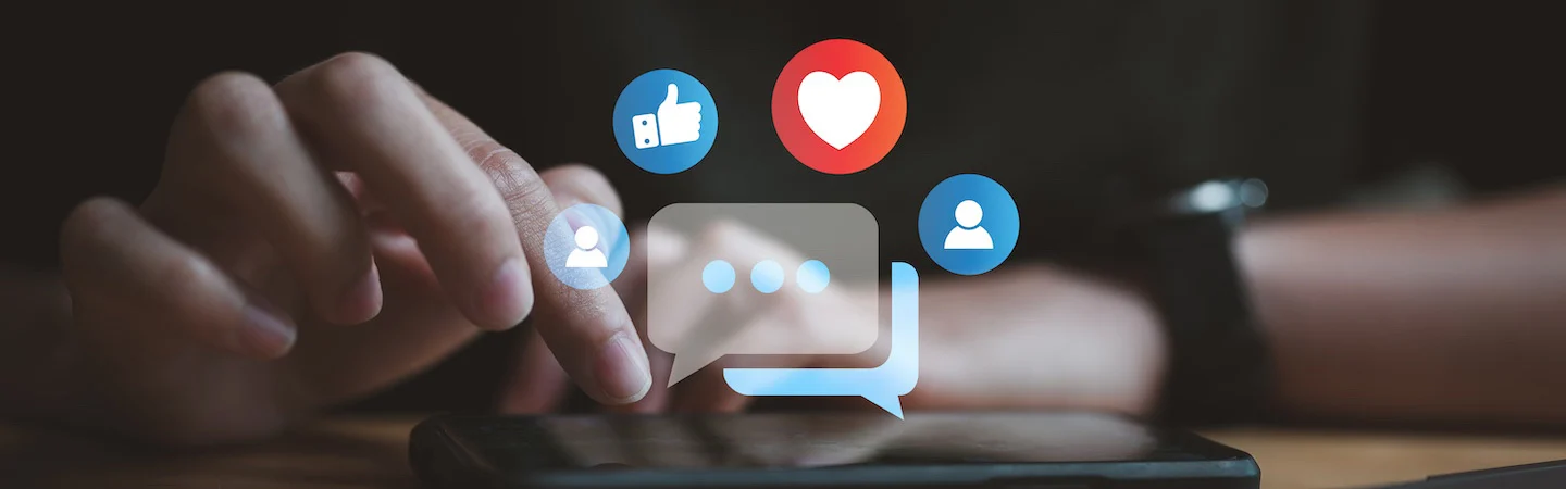 Close-up of a hand touching a smartphone screen with social media icons like thumbs up, heart, and chat bubbles floating above.