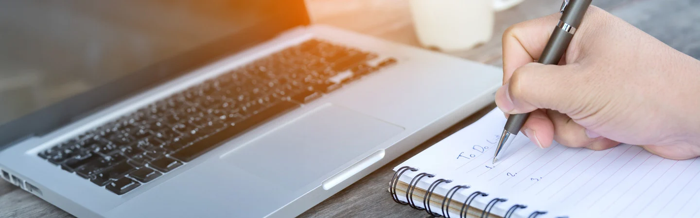 Close-up of a hand writing in a spiral notebook with a pen, next to an open laptop on a wooden surface.