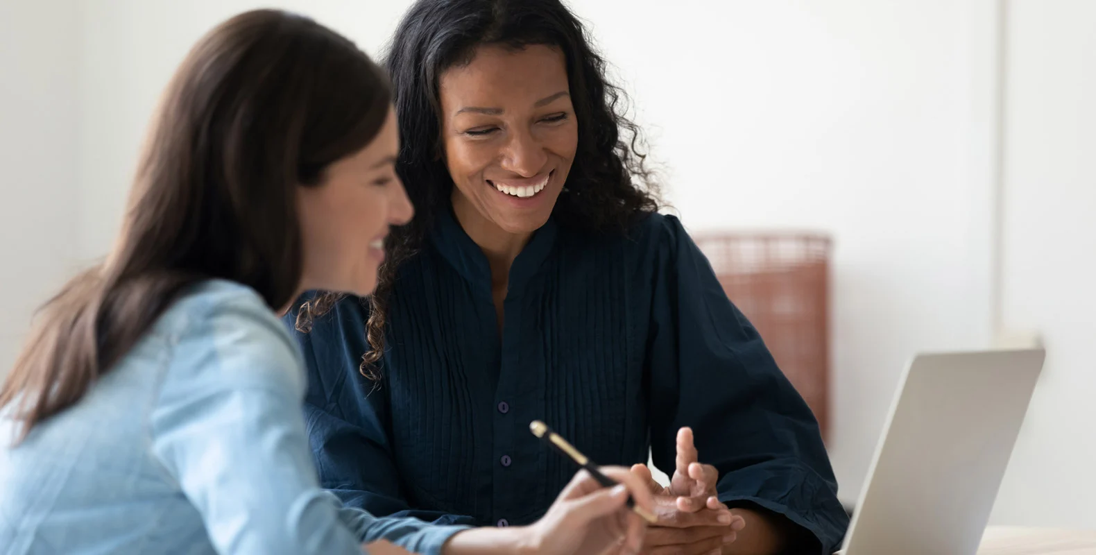 Two women sitting at a table, smiling and discussing something while looking at a laptop screen.