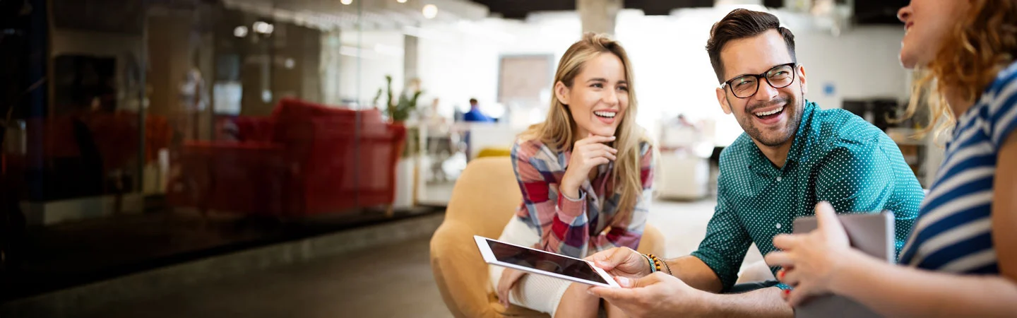 Three people sit and laugh together in a modern office space, one holding a tablet, engaging in a lively conversation.