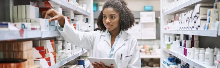 A pharmacist in a white coat holds a tablet and organizes medication on shelves in a pharmacy.