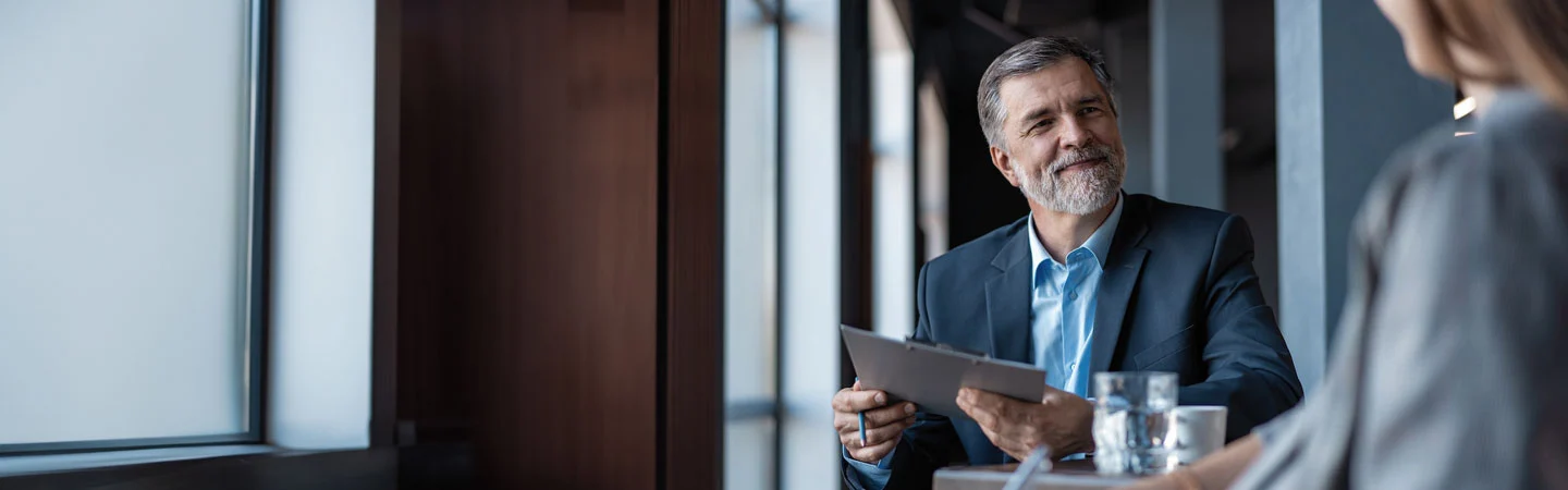 A man in a suit smiles, holding a tablet, sitting at a table with a blurred person in the foreground.