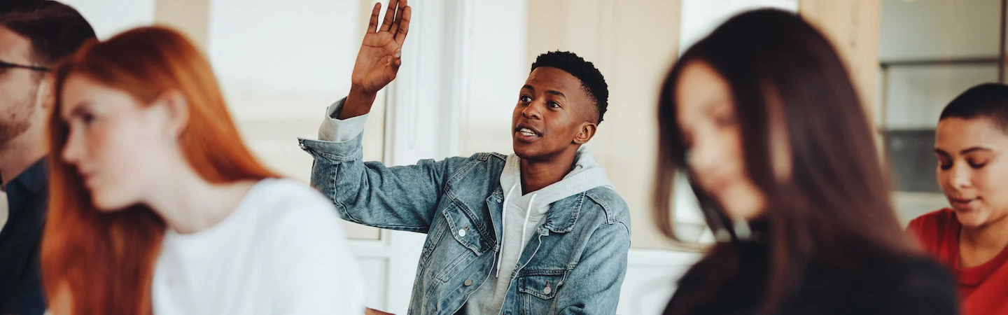 A student raising his hand in a classroom with other students around him.