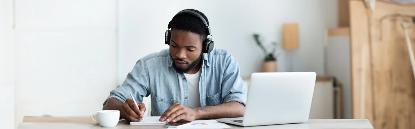 Man wearing headphones, writing in a notebook, seated at a desk with a laptop and a cup of coffee.