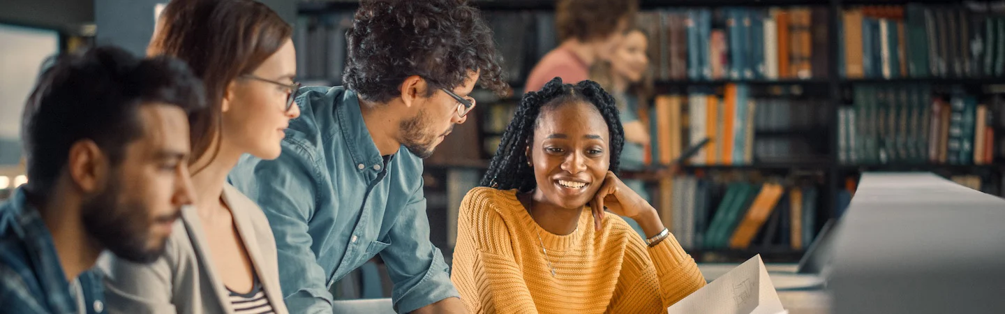 A diverse group of five people engaged in discussion while studying in a library filled with bookshelves.